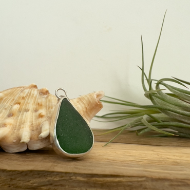 Green seaglass made into a necklace, resting on a shell with an implant in the back of the image.